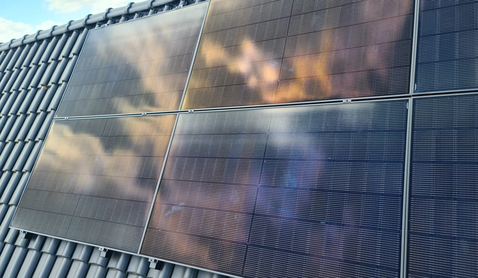 Zwarte zonnepanelen op een pannendak, met wolken weerspiegeld in het glas
