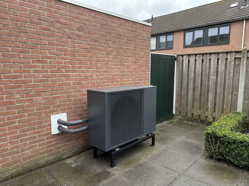 Antraciet buitenunit van een warmtepomp in een achtertuin, tegen de gevel