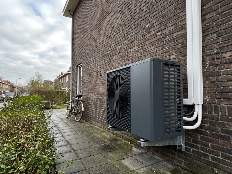 Buitenunit van een warmtepomp aan een muurbeugel langs een zijpad